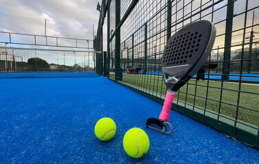 A padel or paddle tennis racket and a bright yellow tennis ball lie on a blue court. The net and a...