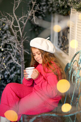 A little girl in a festive winter setting, sitting on a vintage bench and holding a warm cup. Child wearing a bright pink cozy outfit and a white beret. Christmas decorations. Holidays New Year concep
