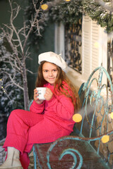 A little girl in a festive winter setting, sitting on a vintage bench and holding a warm cup. Child wearing a bright pink cozy outfit and a white beret. Christmas decorations. Holidays New Year concep