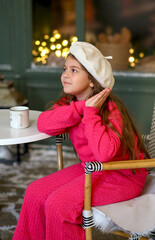 Adorable little girl in a festive winter setting, sitting on a vintage bench and holding a warm cup. Child wearing a bright pink cozy outfit and a white beret, smiling gently. New Year concept