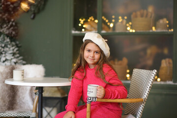 Adorable little girl in a festive winter setting, sitting on a vintage bench and holding a warm cup. Child wearing a bright pink cozy outfit and a white beret, smiling gently at the camera. Christmas