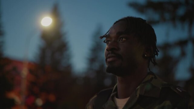 Black Soldier Under Streetlight At Dusk, Wearing Camouflage Jacket And Backpack, Pine Trees And Warm Bokeh Lights In Background, Closeup Portrait With Contemplative Expression, Cinematic Shallow
