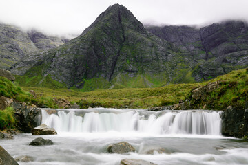 Fototapeta premium Waterfall and water stream, centered steep rocky mountain and ridge covered by clouds in background