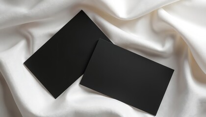 Two blank black cards lay on elegant white satin fabric. Minimal setup perfect for branding mockups business stationery. Use for presentations, advertising, identity design projects.