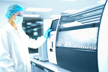 Caucasian female scientist in full safety gear operating automated analyzer in sterile lab....