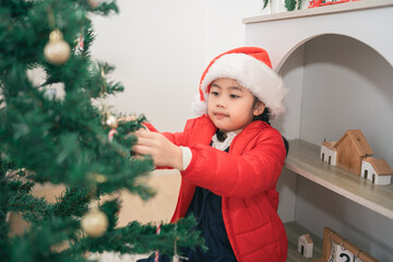 Little Girl in Red Jacket and Santa Hat Decorates Christmas Tree with Ornaments, Festive Atmosphere in Cozy Holiday Setting