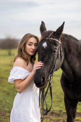 Obraz premium Elegant woman in white off-shoulder dress with a calm black horse in a sunny countryside field