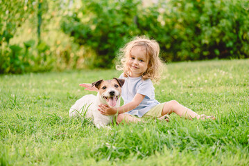 Happy small girl hugging jack russell terrier dog on green grass lawn - child pet friendship