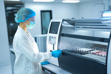 Naklejka premium Caucasian female scientist in full safety gear operating automated analyzer in sterile lab. Professional researcher setting machine parameters for medical biotechnology experiment.