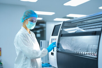 Caucasian female scientist in full safety gear operating automated analyzer in sterile lab. Professional researcher setting machine parameters for medical biotechnology experiment. © ultramansk