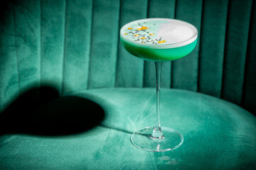 A vibrant, bright green cocktail with white foam, garnished with yellow sprinkles and black seeds, served in an elegant coupe glass on a plush emerald velvet chair.