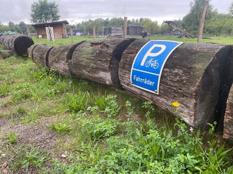 Fahrrad Parkplatz f&uuml;r G&auml;ste, markiert durch ein blaues Parkschild auf gestapelten Baumst&auml;mmen in l&auml;ndlicher Umgebung. Das Motiv steht f&uuml;r einfache Infrastruktur, Orientierung , Alltag im Au&szlig;enbereich.