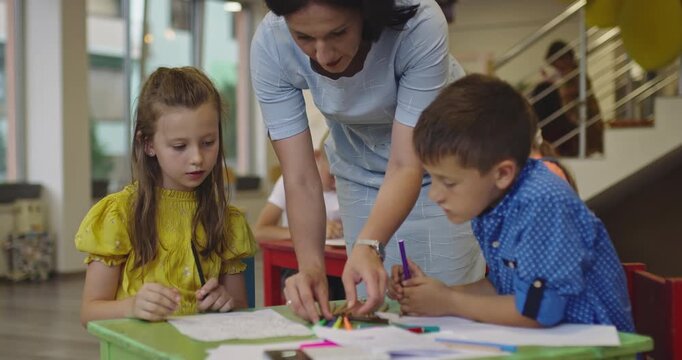 Creative kids during an art class in a daycare center or elementary school classroom drawing with female teacher. 