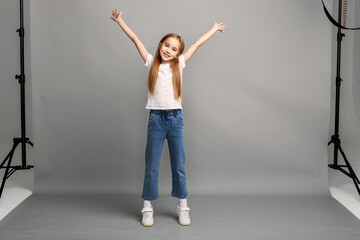 Happy young caucasian girl with long hair and arms raised celebrating success or joy. Kid posing for professional studio photoshoot on gray background. Fashion model in jeans and white T-shirt