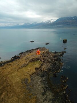 Calm coastal scene with rocky shoreline, red and orange traditional Icelandic lighthouse, and distant snow-capped mountains under cloudy skies, evoking peaceful solitude.