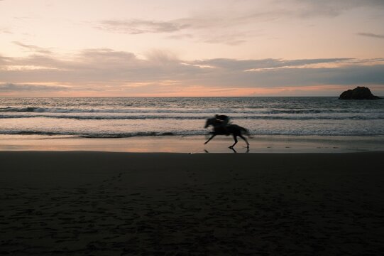 Blurred silhouette of a person running on a beach at sunset with calm ocean waves and a distant rock formation, evoking freedom and nature's beauty.