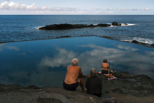 Three people sitting on rocky shore beside a calm tidal pool reflecting the sky, overlooking the ocean and distant horizon under a clear sky.