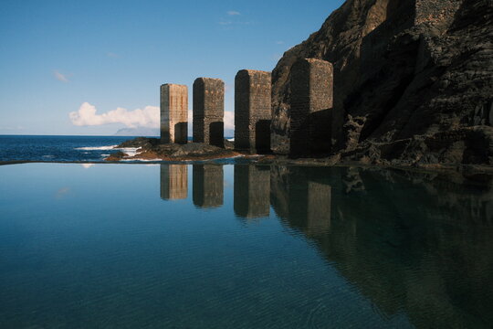 Ancient stone pillars reflected in calm water near a rocky coastline under a clear blue sky, creating a peaceful and historic atmosphere. - Powered by Adobe