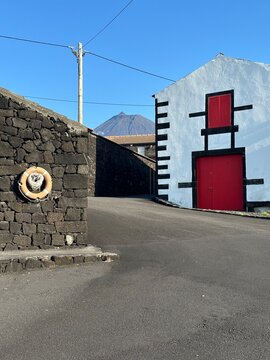 A rustic street scene featuring traditional stone walls and a white building with red doors and shutters under a clear blue sky, evoking a serene village atmosphere.