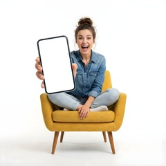 Excited young woman sitting on yellow armchair showing big smartphone with blank white screen isolated on white background.