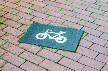 A clear white bicycle icon is stenciled onto a dark rectangular patch of asphalt, which is set into a textured brick walkway with small green grass sprouts emerging from the gaps