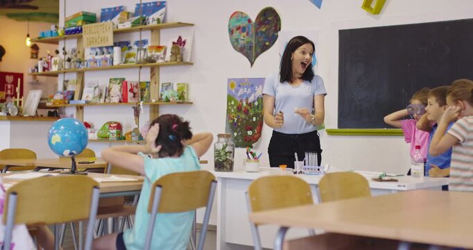 Smiling teacher conducting a science experiment with a balloon and lighter for excited elementary students in a classroom, while kids cover their ears in anticipation of the pop
