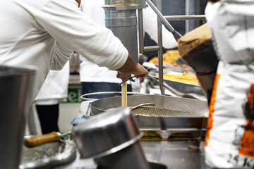 Cook operates a churro maker over hot oil in a busy kitchen; selective focus documents the process, heat and stainless textures of street‑food preparation.