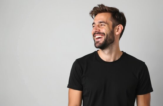 Man with beard laughs outdoors against bright backdrop. He wears a black t-shirt and looks to the side with a wide smile, showing happiness. He has dark hair, styled casually.