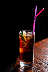 A tall glass with a dark drink, ice cubes, and lime slices on a textured wooden table