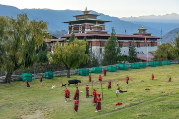 Paro, Bhutan - 26 November 2025: View of monks in vibrant red robes playing football on the green field near Rinpung Dzong, with mountains in the distance.