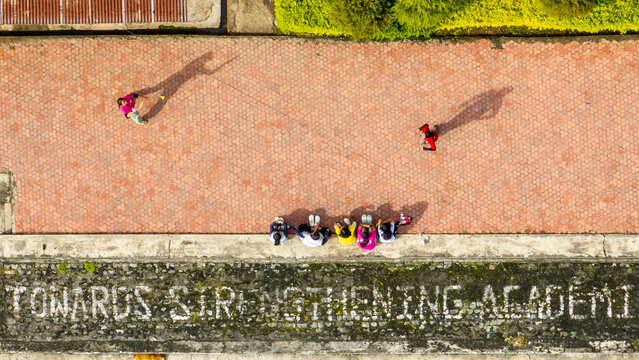 Aerial view of students gathered near a wall with faded inscriptions, their shadows stretching across the brick path, Punakha Central School, Punakha, bhutan.