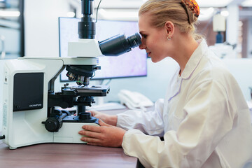 Young Caucasian female scientist adjusting microscope in modern laboratory. Researcher analyzing histology slide displayed on background monitor for medical diagnosis.