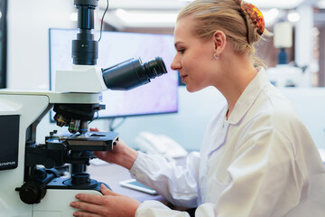 Young Caucasian female scientist adjusting microscope in modern laboratory. Researcher analyzing histology slide displayed on background monitor for medical diagnosis.