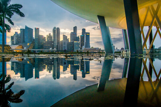 View of the city skyline shimmering in mirrored reflections on the water, captured beneath modern architecture, as twilight paints the sky, Raffles Place and Marina Boulevard, Singapore.