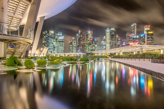 View of a tranquil pond reflecting the vibrant skyline under a cloudy night sky, with ArtScience Museum framing the scene, Raffles Place and Marina Boulevard, Singapore.