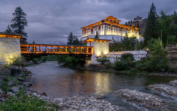 View of the illuminated Rinpung Dzong fortress glows majestically above the flowing river and connected bridge, Rinpung Dzong, Paro, Bhutan.