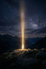 night landscape featuring a glowing luminous figure standing on a rocky mountain peak with a powerful beam of light shooting into the starry milky way galaxy.