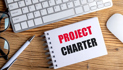 Notebook with text project charter on wooden desk with keyboard, pen, and glasses, symbolizing...