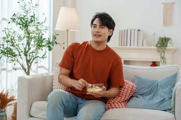 young Asian man in a t-shirt and jeans is sitting comfortably on a light colored sofa in a bright, modern living room, watching TV and holding a small bowl of popcorn or snack.