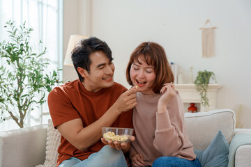 Happy young Asian couple eating popcorn together, man feeding woman in bright living room. They are relaxing and bonding while sitting on the sofa at home.