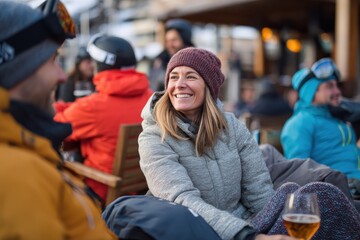 A cheerful woman in a winter jacket and knit hat smiles while holding a drink at a busy apr&egrave;s-ski terrace with other skiers relaxing in the snowy alpine resort environment