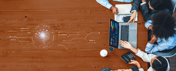 A diverse business team collaborates around a workstation, focused on data analysis displayed on a laptop. They engage in strategic planning, surrounded by documents and coffee. Trope