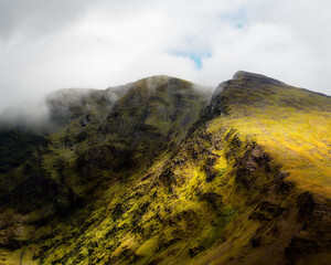 This scenic shot showcases the rugged terrain of the Coomloughra Horseshoe Mountains. Green grass covers the slopes and hiking trails are clearly visible. Clouds partially obscure the mountain peaks.