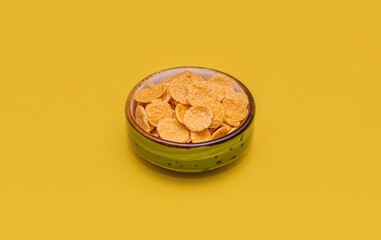 A vibrant shot of golden crunchy cornflakes cereal filling a decorative mini bowl. Cornflakes isolated against a bright yellow background. Use for food marketing.