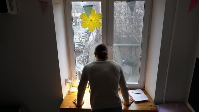 High angle back view of a person leaning over a wooden desk and looking out of a window in a home office, with soft natural light illuminating the scene and papers scattered on the workspace
