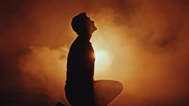 Dramatic silhouette of a man kneeling in swirling orange smoke. Person raising head from defeat to hope. Mental health and prayer concept