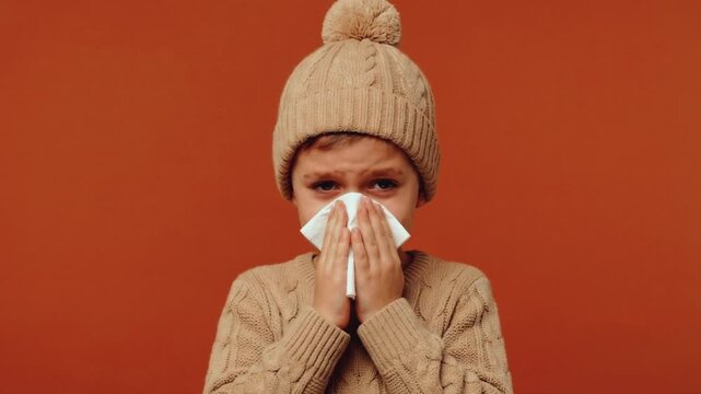 Sick child blowing nose into a tissue on orange background. Boy with flu or allergy symptoms wearing a beige sweater and beanie hat