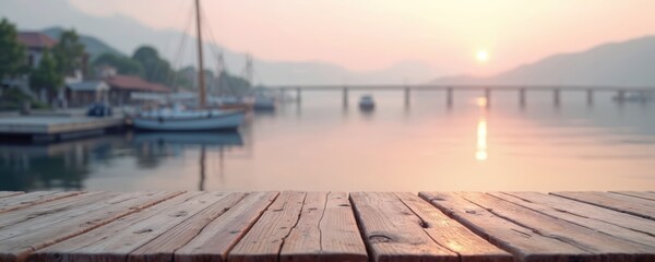 Wooden tabletop surface with blurred lake harbor and sunset backdrop. Boats dock near mountains across calm water reflecting pink sky. Peaceful waterscape scene perfect for product placement.