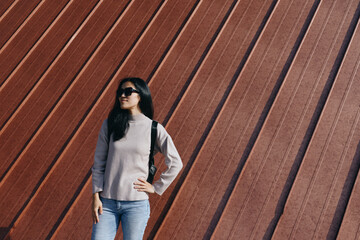 Woman wearing sunglasses, denim jeans and a sweater, with a backpack, standing confidently against...