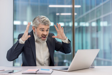 Mature businessman experiencing intense stress and frustration, yelling at a laptop screen in an...